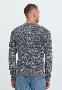 Vyras su trumpais plaukais, apsivilkęs juodai-baltu dryžuotu megztiniu ir mėlynomis džinsomis, stovi atsukęs nugarą prieš paprastą šviesią foną.