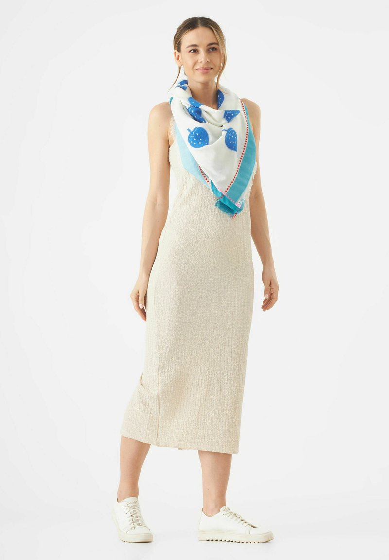 Beige midi-jurk van gestructureerde stof, geaccentueerd met een blauw-witte sjaal met aardbeitjespatronen en een levendige rand. Witte sneakers maken de look af.