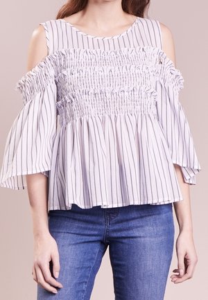 Frau trägt eine weiße Cold-Shoulder-Bluse mit schwarzen vertikalen Streifen und gerafften Details, kombiniert mit blauen Jeans.