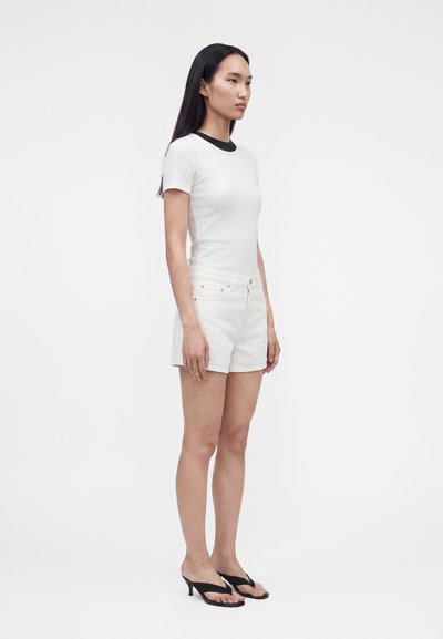 Γυναίκα που στέκεται πλάγια, φοράει ένα στενό λευκό μπλουζάκι, λευκά σορτς και μαύρα σανδάλια με τακούνι σε απλό λευκό φόντο.