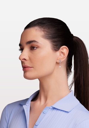 Cercei argintii în formă de clemă de siguranță, cu un mic dejun de perlă. Modelul este prezentat în profil, purtând o cămașă cu guler de culoare albastră deschisă.