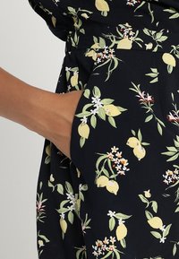 Svart tyg med citron- och blommönster; en hand sträcker sig in i en sidoficka och visar den avslappnade designen och det lätta materialet.