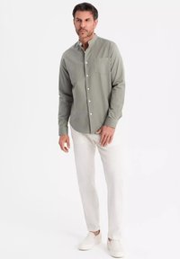 Camicia verde chiaro con bottoni e tasca sul petto, abbinata a pantaloni bianchi e scarpe slip-on bianche. Il tessuto appare liscio e informale.