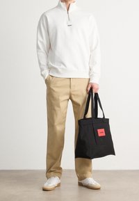 Felpa bianca con zip e logo nero, pantaloni beige, scarpe da ginnastica bianche e una borsa nera con etichetta rossa. Abbigliamento casual.