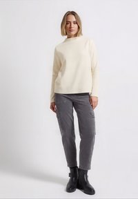 Kremowa bluza z okrągłym dekoltem, połączona z szarymi spodniami typu cargo z bocznymi kieszeniami oraz czarnymi botkami za kostkę. Ogólnie gładka faktura.