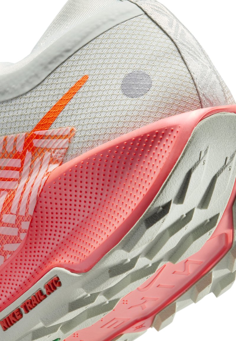 Zapatilla deportiva gris con un acabado texturizado, detalles en naranja brillante, un talón rosa y una suela resistente con surcos definidos, además de la marca Nike Trail ATC.