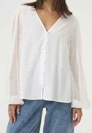 Vrouw die een witte blouse draagt met een getextureerde knoopsluiting aan de voorkant en doorschijnende, patroonlange mouwen, gecombineerd met blauwe denim jeans.