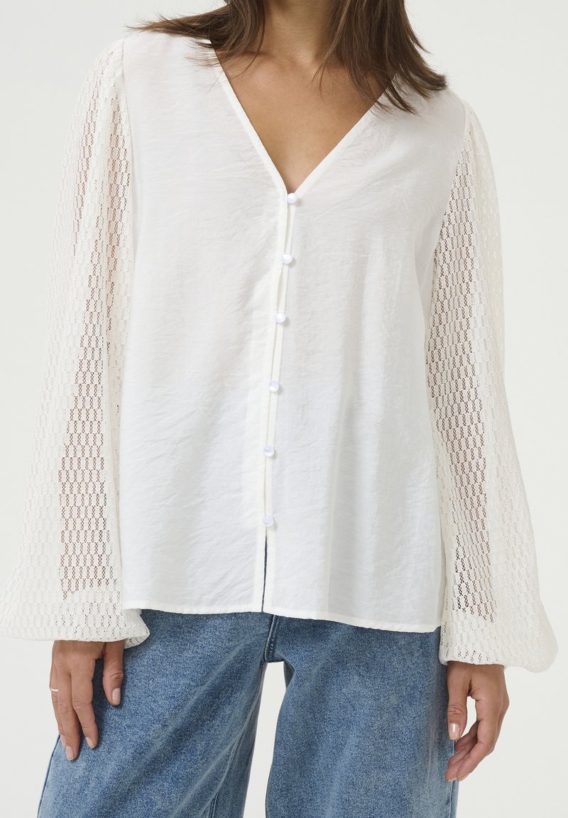 Femme portant un chemisier blanc avec un devant boutonné texturé et des manches longues transparentes à motifs, associé à un jean en denim bleu.