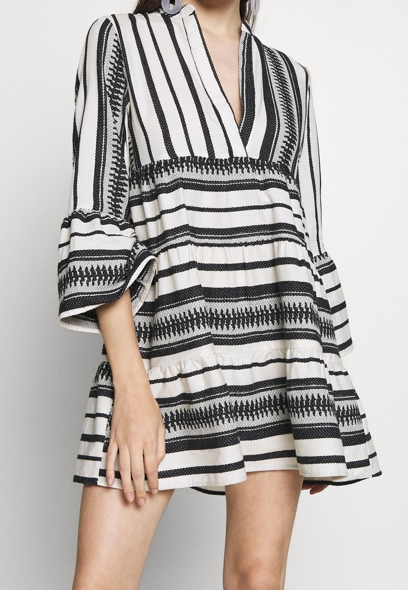 Robe rayée en blanc cassé et noir, avec un col en V, des manches longues évasées, une taille froncée et un ourlet à volants avec des accents texturés.