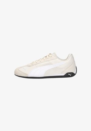 Lichtbeige Puma sneaker met witte streepaccenten, geperforeerde zijkanten, veterluiting en zwarte zool met Puma-logo op de hiel.