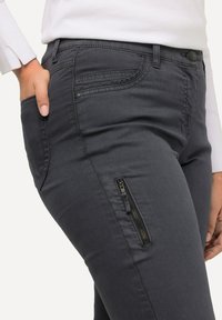 Graue Stretchleggings mit schmaler Passform, featuring Fronttaschen, dezenten Nähten und einer seitlichen Reißverschlusstasche mit goldenen Reißverschlussdetails.