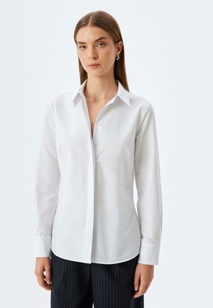 Camicia bianca con colletto, maniche lunghe e texture liscia; presenta una patta frontale e orlo dritto, abbinata a pantaloni a righe.