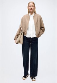 Beige bomberjakke over en hvit skjorte, kombinert med mørke vide jeans og svarte treningssko med hvite detaljer. Bærer en beige handlenett.