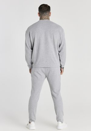 Mand med hals tatovering iført en grå sweatshirt og joggingbukser, stående med ryggen mod kameraet på en ensfarvet lys baggrund.