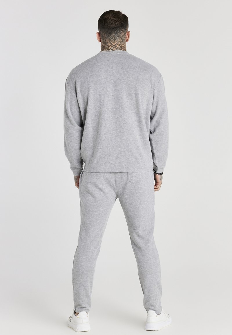 Homme avec un tatouage au cou portant un sweat-shirt gris et un pantalon de survêtement, debout de dos sur un fond clair uni.