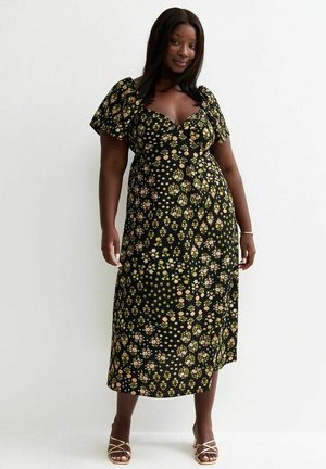 Kobieta ubrana w czarną sukienkę midi z bufkami i małym żółtym wzorem kwiatowym, stojąca na tle jednolitego białego tła.