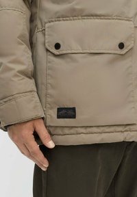 Beige isolierte Jacke mit einer Vorderseite Tasche, schwarzen Druckknöpfen und einem Etikett am Saum. Hand ruht auf der Tasche. Texturierter Stoff.