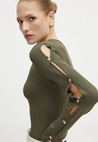 Bodysuit verde oliva con dettagli cut-out sulle spalle e accenti in bottone. Seduta aderente con superficie liscia, dotato di maniche lunghe.