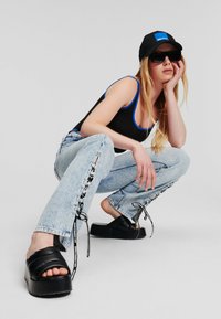 Zwarte platform sandalen, zwart tanktop met blauwe randen, en licht gewassen jeans met veters aan de zijkanten. Model zit in squat, draagt een zonnebril en een pet.
