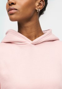 Sweat à capuche rose clair avec un col côtelé et un tissu doux. Il présente une coupe décontractée et une capuche avec des plis superposés. Aucun motif ni accent.