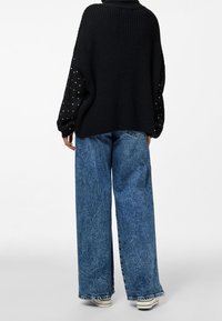 Svart stickad tröja med strukturerat mönster och pärldetaljer på ärmarna, kombinerad med blå vida jeans med fransig fåll.