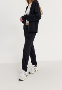 Schwarzer Kapuzenpullover mit Reißverschluss, kombiniert mit passenden schwarzen Jogginghosen. Auffällige Merkmale sind eine lockere Passform, ein elastischer Bund und weiße Turnschuhe.