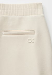 Beige sweatpants with a ribbed waistband, featuring a back pocket and embossed logo detail. Soft texture with a smooth finish.
