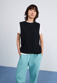 Débardeur noir texturé sans manches avec un motif en grille superposé sur un t-shirt blanc, associé à un pantalon de jogging bleu clair avec un logo.