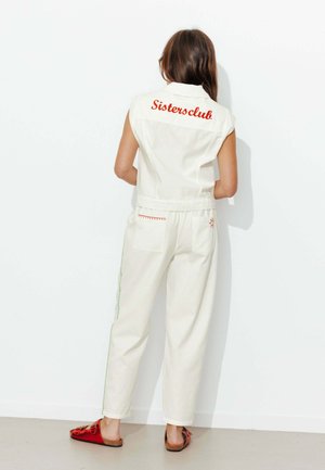 Femme portant un gilet blanc sans manches avec « Sistersclub » en rouge dans le dos, un pantalon blanc avec des coutures rouges et vertes, et des chaussures rouges à enfiler.