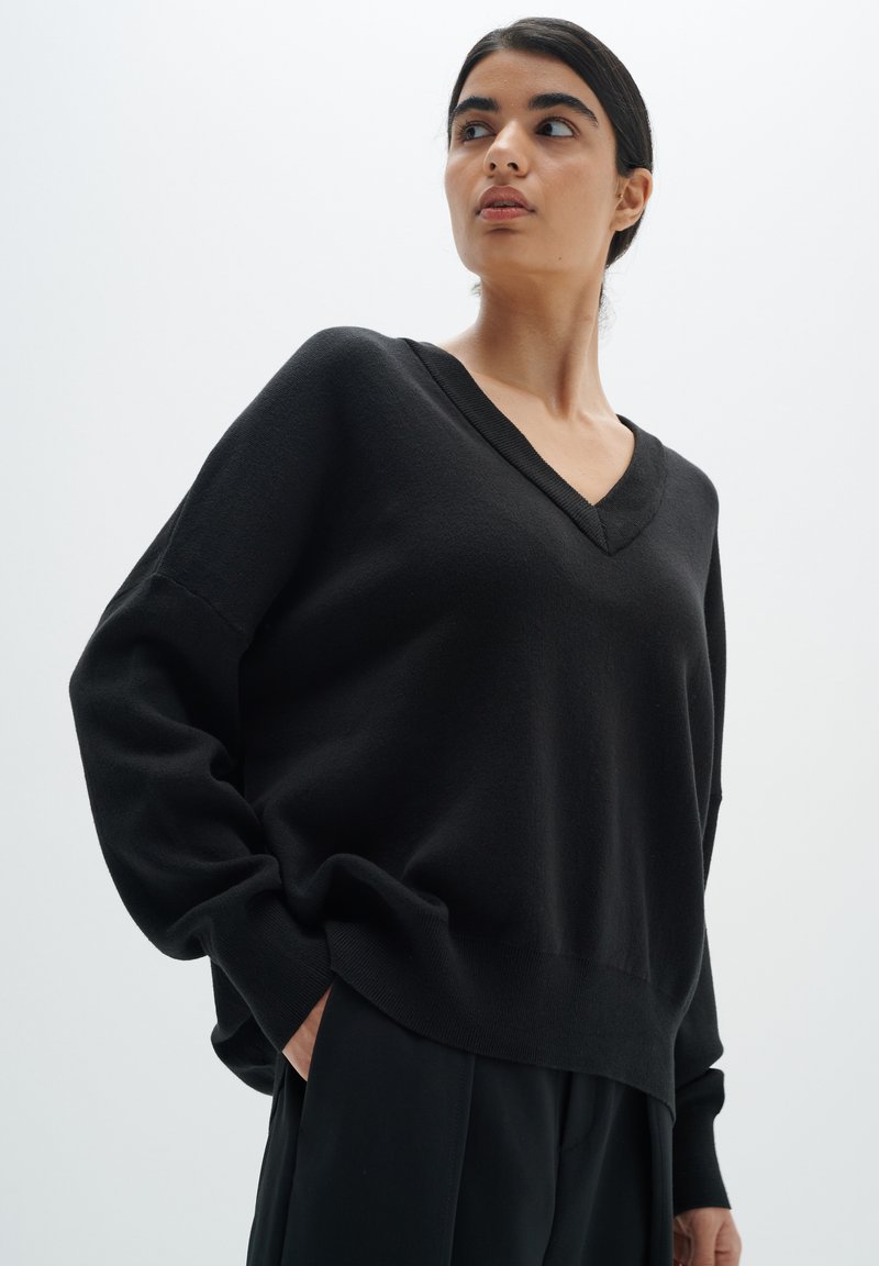 Black knit sweater with a V-neck, dropped shoulders, and a relaxed fit. Soft texture and ribbed cuffs, paired with tailored black trousers.