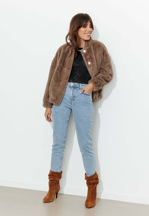 Femme portant une veste en fausse fourrure marron, un haut noir, un jean taille haute bleu clair et des bottes en daim marron à talons, debout contre un mur blanc.