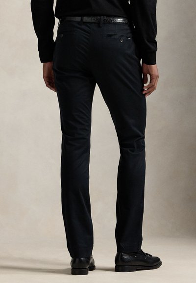 Muž oblečený v čiernych slim-fit nohaviciach, čiernom koženom opasku, čiernych spoločenských topánkach a čiernej košeli s dlhým rukávom, stojaci pred jednoduchým pozadím.