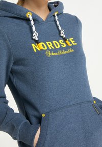 Modrá mikina s kapucí, bílými a černými šňůrkami, žlutým nápisem "NORDSEE Schmuuddelwedda" a rukou osoby ve přední kapse.