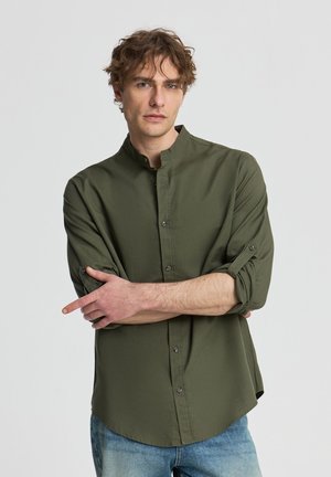 Giovane uomo con capelli ricci castani che indossa una camicia verde oliva con bottoni e maniche arrotolate e jeans blu, in piedi con le braccia incrociate.