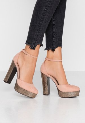 Lyserøde fløjlsplatformsandaler med en guldmønstret sål og en kraftig hæl. Ankelstrop med en cirkulær spænde detalje.