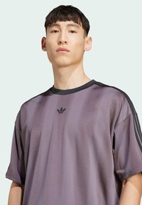 Fialové pruhované sportovní tričko s kulatým výstřihem, černými akcenty a logem na hrudi. Vyrobeno z lehkého materiálu s texturovaným povrchem.