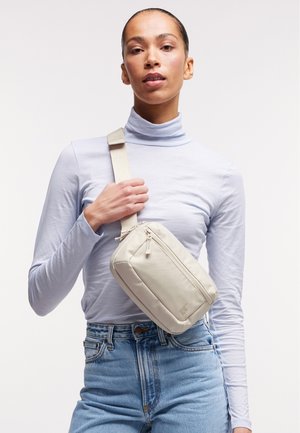 Femme portant un col roulé bleu clair et un jean, tenant un sac bandoulière beige croisé sur sa poitrine sur un fond blanc uni.