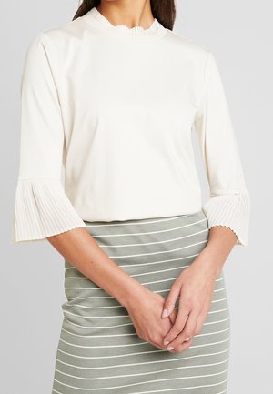 Femme portant un chemisier blanc plissé à manches évasées, associé à une jupe à rayures horizontales vertes et blanches, les mains détendues jointes devant.