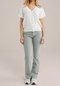 Žena v bílém krátkém rukávu zapnutém svetříku, světle šedých přiléhavých kalhotách a bílých teniskách stojí na jednolité béžové pozadí.