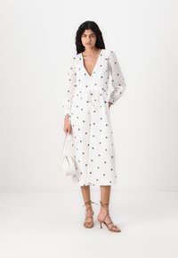 Vit midiklänning med svarta blommönster, V-ringning, långa puffärmar och insvängd midja. Matchas med sandaler med remmar och en handväska.