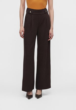 Femme portant un pantalon large taille haute marron foncé avec des boutons dorés et des chaussures brunes à bouts pointus, debout devant un fond uni.