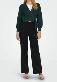 Dunkelgrüne, gestrickte Wickeljacke mit langen Ärmeln und Bindedetail, kombiniert mit schwarzen, hochtaillierten Hosen mit weitem Bein und schwarzen Sandalen.