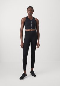 Top desportivo preto com fecho na frente e detalhe do logótipo, combinado com leggings pretas de cintura alta e sapatilhas pretas.