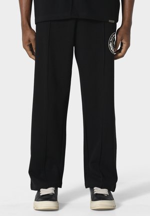 Person iført løse sorte bukser med hvidt logo på højre lår og sorte sneakers med hvide såler og snørebånd, stående mod en ensfarvet baggrund.