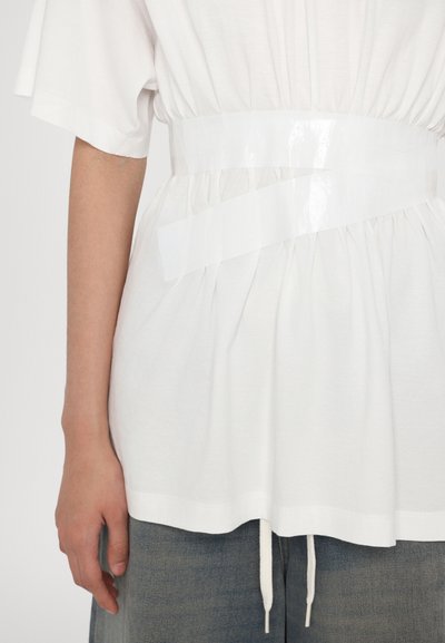Vit kortärmad skjorta med en rynkdesign och ett skinnande transparent plastbälte som är snurrat runt midjan. Sömmen har en dragsko.