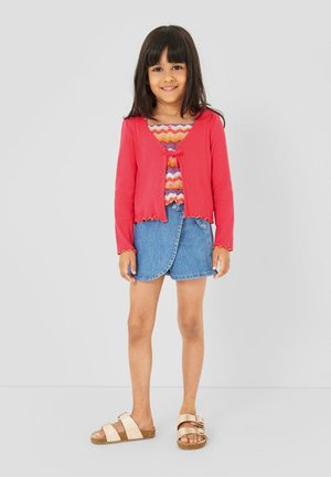 Fille souriante, portant un cardigan rouge, un haut à motifs zigzag colorés, une jupe en jean et des sandales beige, debout devant un fond gris clair uni.