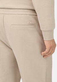 Béžové technické joggery s textúrovaným povrchom, s zadnou kapsou a malým značkovým štítkom. Sú prítomné viditeľné švy.