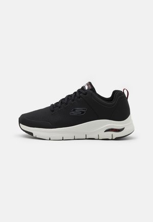 Baskets de sport noires avec une tige en maille, surface texturée et semelle intermédiaire blanche. Présente un design à lacets et un logo sur le côté, avec un accent rouge.