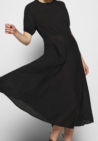 Vestido negro con un escote redondo, mangas cortas abullonadas y una silueta en A fluida. La tela texturizada crea sutiles ondulaciones.