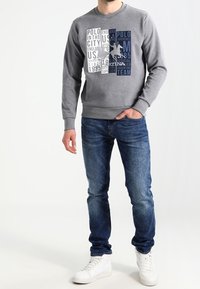 Šedá polokošile s kulatým výstřihem, zdobená grafickými bloky textu v námořnické modři a bílé. V kombinaci s modrými džíny a bílými teniskami. Casual střih.